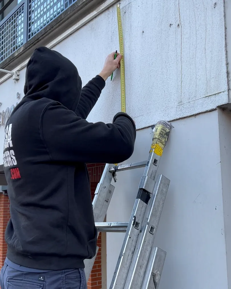 Un poseur prend les mesures d'une enseigne sur une façade, échelle en place